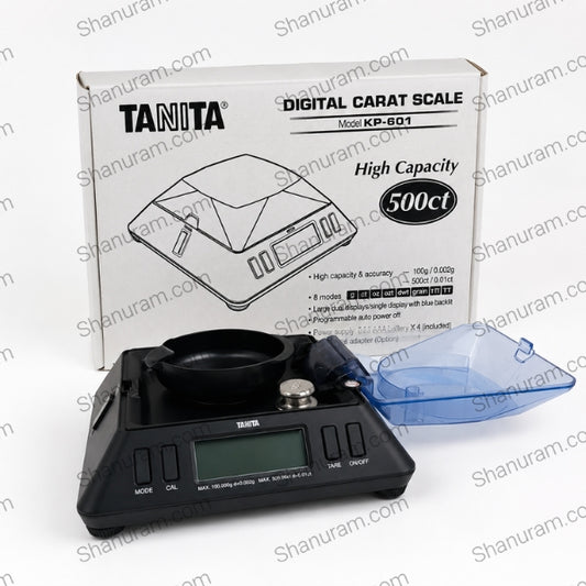 Tanita digital carat scale model KP-601 shown with its retail box and a translucent blue lid on a white background. The scale is black with a digital display screen, and the packaging box is white with black text and line drawings.
